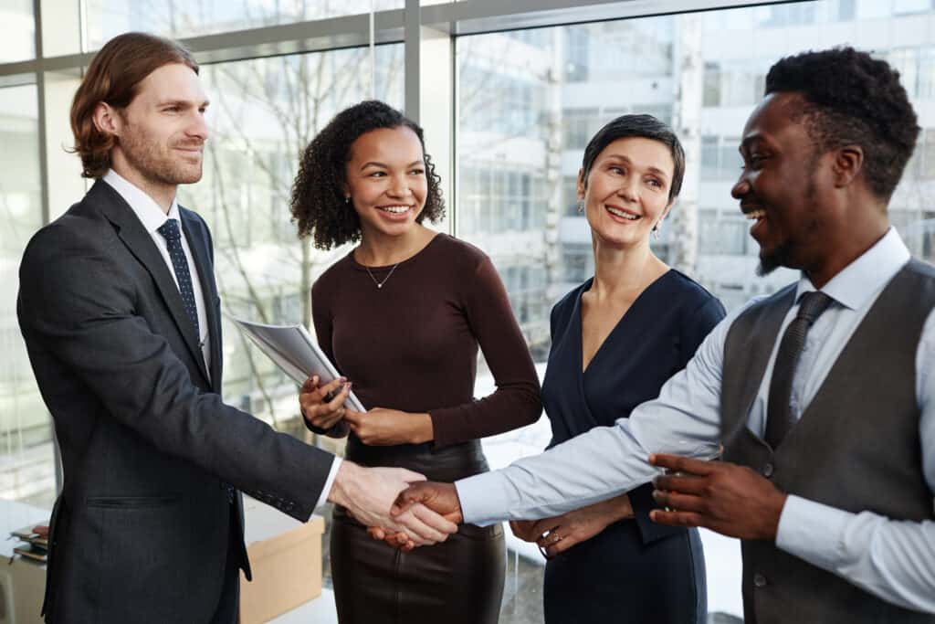 Smiling business partners shaking hands in office standing with drinks